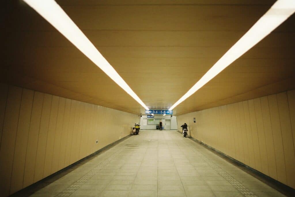 A long pedestrian tunnel with bright overhead lights and people on scooters